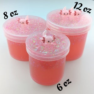 May include: Three clear plastic containers of pink slime with glitter. The containers are labeled 8 oz, 6 oz, and 12 oz. Each container has a pink axolotl charm on top.