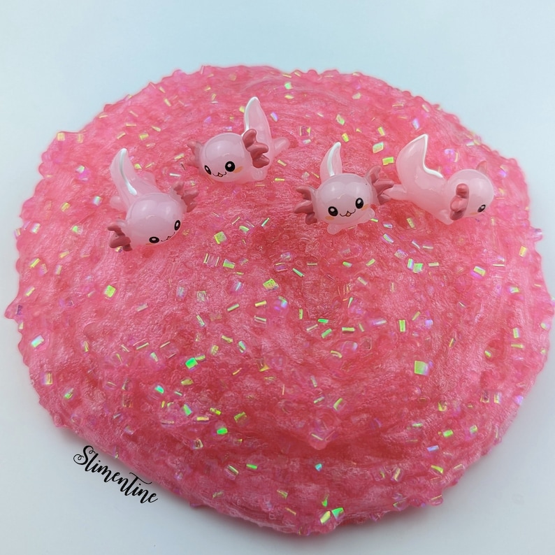 May include: Pink slime with iridescent glitter and four small pink axolotl charms. The slime is in a bowl and the charms are on top of the slime.