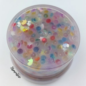 May include: A clear plastic container filled with colorful, translucent, and iridescent beads and glitter. The beads are various sizes and shapes, and the glitter is scattered throughout the container.