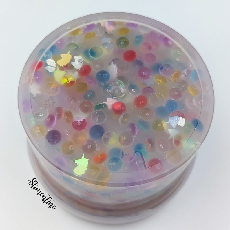 May include: A clear plastic container filled with colorful, translucent, and iridescent beads and glitter. The beads are various sizes and shapes, and the glitter is scattered throughout the container.