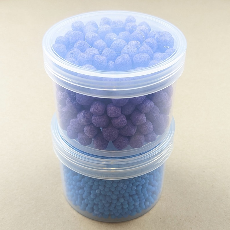 May include: Two clear plastic containers stacked on top of each other. The top container is filled with small, round, purple candies. The bottom container is filled with small, round, blue candies.