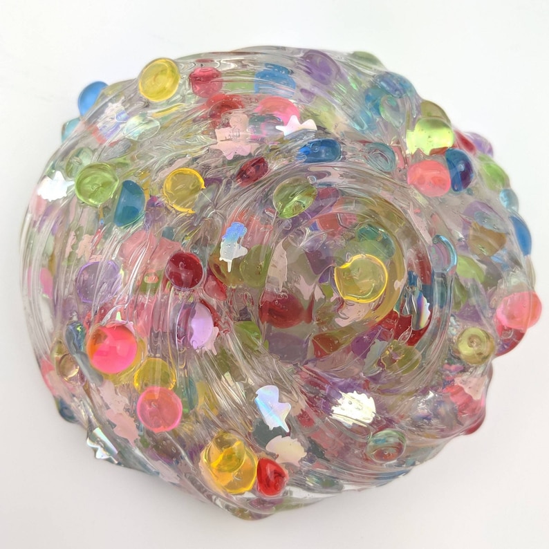 May include: Clear slime with colorful beads and glitter. The slime is in a round shape and has a smooth, glossy texture.