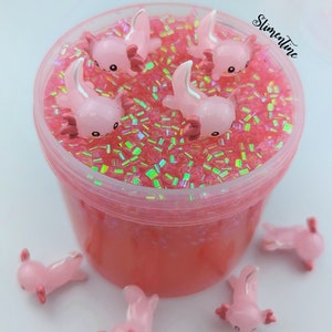May include: Pink slime with iridescent glitter and pink axolotl charms. The slime is in a clear plastic container with a pink lid. There are additional axolotl charms outside of the container.