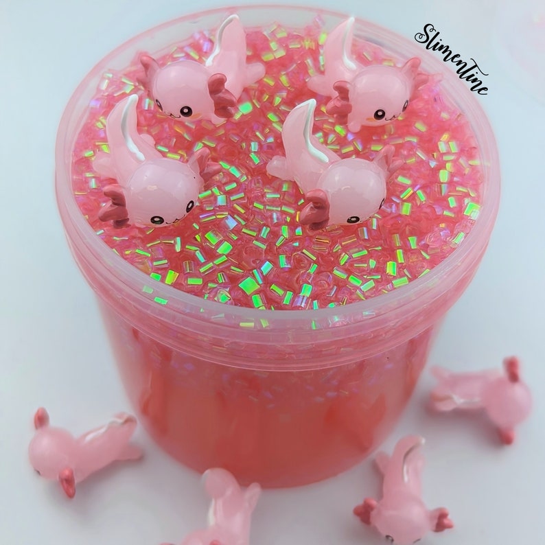 May include: Pink slime with iridescent glitter and pink axolotl charms. The slime is in a clear plastic container with a pink lid. There are additional axolotl charms outside of the container.