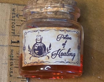 May include: A small glass jar with a cork stopper, filled with a reddish liquid. The jar has a label that reads "Potion of Healing" with a graphic of a potion bottle. The jar is sitting on a wooden surface.
