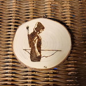 May include: A wood-burned image of a hooded figure with a bow and arrow, on a round wooden plaque.
