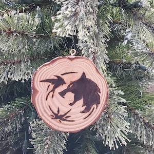 May include: A wooden Christmas ornament with a burnt-in design of a wolf's head and a stylized wolf paw print. The ornament is hanging on a green Christmas tree.