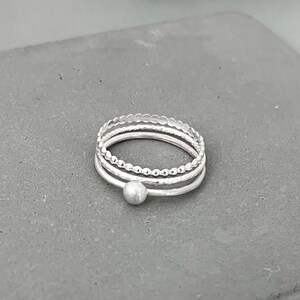 Puede incluir: Un conjunto de cuatro anillos apilables de plata con diferentes texturas. Los anillos están apilados uno encima del otro y tienen una pequeña bola de plata en la parte inferior.