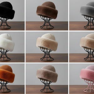 Könnte beinhalten: Eine Auswahl an Angora Strick Hüten in verschiedenen Farben, darunter Schwarz, Braun, Grau, Weiß, Beige, Orange und Rosa. Jeder Hut hat eine gerippte Strickkrone und einen flauschigen Kunstfellrand. Die Hüte werden auf dekorativen Metallständern präsentiert.