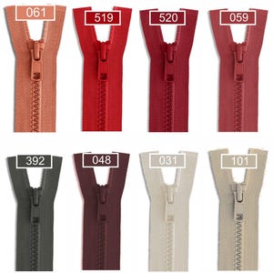 May include: A collection of twelve plastic zippers arranged in two rows, featuring a variety of colors including shades of pink, red, burgundy, grey, brown, and beige. Each zipper is labeled with a specific numerical code: 213, 061, 519, 520, 059, 527, 850, 392, 048, 031, 101, and 551.