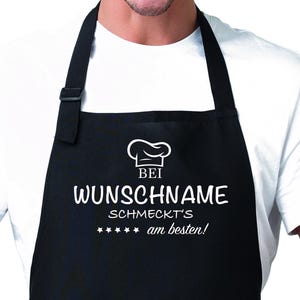 Könnte beinhalten: Schwarze Schürze mit weißem Text, der "Bei WUNSCHNAME schmeckt's am besten!" lautet, mit einem Kochmützen-Symbol über dem Text.