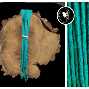 Könnte beinhalten: Ein Bündel türkisfarbener synthetischer Dreadlocks, etwa 50 cm lang, mit einem silbernen Ring oben. Die Dreadlocks werden auf einer Holzoberfläche gezeigt.