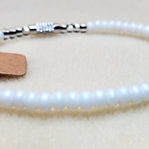 May include: White beaded bracelet with a silver clasp. The bracelet has a brown tag with the word "Omi" written on it.