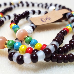 May include: Three beaded bracelets with brown, white, and multi-colored beads. The bracelets are stacked on top of each other. A brown tag with the word "Oni" is attached to the bracelets.
