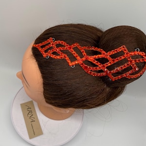 May include: A geometric red rhinestone hair accessory is shown on a mannequin head with brown hair styled in a bun. The accessory is made of small, sparkling red rhinestones. A tag with the name "ARNA" is visible. The hair accessory is a striking piece.