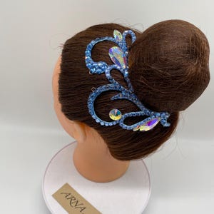 Könnte beinhalten: Ein blaues Haaraccessoire mit schillernden Kristallen und Strasssteinen. Das dekorative Stück ist auf einem Schaufensterkopf mit braunem Haar befestigt, das zu einem Dutt frisiert ist. Das Accessoire hat ein florales Design und ist mit mehreren Kristallen verziert.
