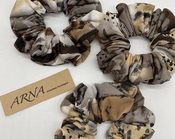 May include: Three fabric hair scrunchies featuring a brown, beige, and black abstract pattern. A rectangular tan paper tag is included with the text ARNA creation boutique printed in black ink on the surface of the tag.