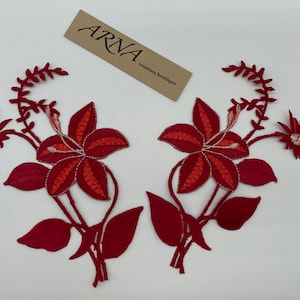 May include: Two red embroidered fabric appliques featuring lily flowers with orange centers and green leaves. The appliques are perfect for adding a touch of floral detail to clothing, bags, or home decor projects.