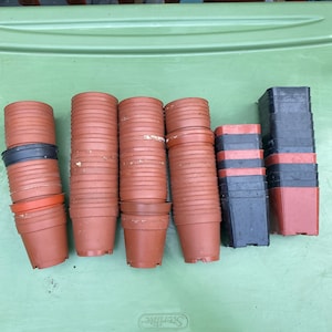 Pode incluir: Uma coleção de vasos de plantas de plástico marrom e preto empilhados em uma superfície verde. Os vasos são de vários tamanhos e estão prontos para plantar.