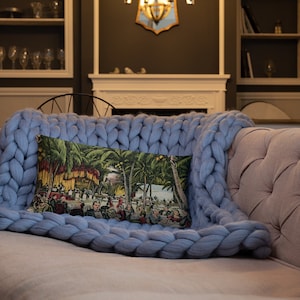 May include: A blue knitted throw blanket with a pillow featuring a vintage illustration of people gathered under palm trees. The pillow has a yellow, red, and green color scheme.