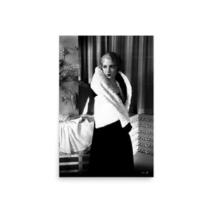 May include: A black and white photograph featuring a person with short, wavy hair wearing a dark, sleeveless dress paired with a thick, white faux fur stole draped over the shoulders. The person is adorned with a multi-strand beaded necklace.