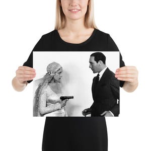 May include: A black and white print featuring a woman in a lace wedding dress and veil holding a small handgun, facing a man in a dark pinstriped suit and tie. The scene captures a dramatic interaction between the two figures in formal attire.