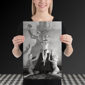 May include: A black and white photographic print featuring a central figure seated on a draped pedestal, wearing an elaborate headpiece and holding a goblet. Two figures kneel on either side, wearing hats and reaching toward the central figure.