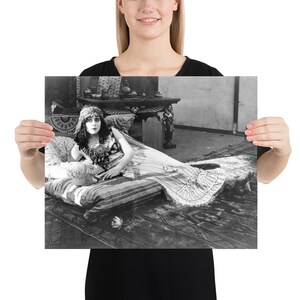 Pode incluir: Uma fotografia a preto e branco de uma mulher em traje deitado numa cama com uma manta estampada. Ela está usando um chapéu e uma túnica longa e fluida. O quarto está decorado com móveis e tapetes ornamentados.
