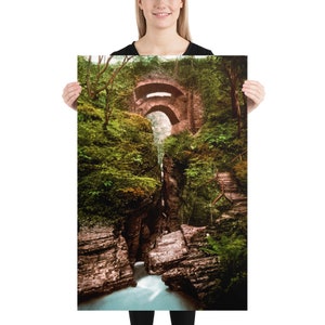 Könnte beinhalten: Eine Steinbogenbrücke über einen schmalen, felsigen Bach in einem üppigen grünen Wald. Die Brücke ist aus braunem Stein gebaut und hat einen abgerundeten Bogen. Der Bach fließt durch eine enge Schlucht, und die Felsen sind mit Moos und Farnen bedeckt.