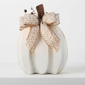 May include: A white decorative pumpkin with a distressed finish, featuring a large beige and white patterned ribbon bow tied at the top. The pumpkin is topped with a dark brown wooden stem and small dark brown decorative berry accents.