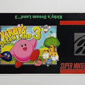 May include: A black Super Nintendo game cartridge with a label for Kirby's Dream Land 3. The label features a pink Kirby character and other characters from the game. The label also includes the Nintendo logo, a "KA" rating, and the text "SNS-AFJE-USA MADE IN JAPAN".