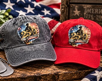 May include: Two baseball caps, one in charcoal grey and one in bright red, each featuring a circular graphic with a soldier silhouette, fireworks, the number 250, and the years 1776 and 2026 printed on the front panels of the hats.