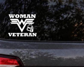 May include: A white vinyl decal applied to a vehicle window featuring the text WOMAN VETERAN in a bold font. Between the words is a stylized letter W with wings and a pair of military-style dog tags hanging from the right side of the letter.