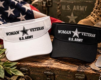May include: Two sun visors, one white and one black, each featuring embroidered text that reads WOMAN VETERAN with a star icon in the center and U.S. ARMY printed below. The visors have a structured brim and a wide headband design.