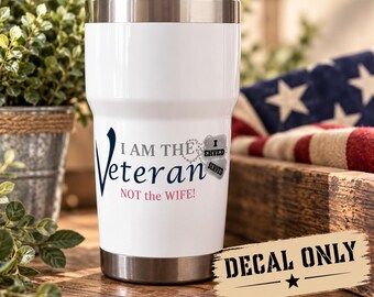 May include: A white stainless steel tumbler with a clear lid featuring the text I AM THE Veteran NOT the WIFE! in dark blue and pink lettering, accompanied by a graphic of two silver dog tags with the words I SERVED.