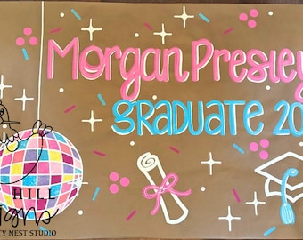 May include: A brown graduation sign with the name "Morgan Presley" in pink and white, and "GRADUATE 2025" in blue and white. The sign includes a disco ball, graduation cap, and diploma illustrations.
