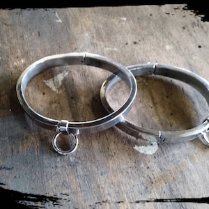 May include: Two silver metal collars with a ring attached to each. The collars are lying on a wooden surface.