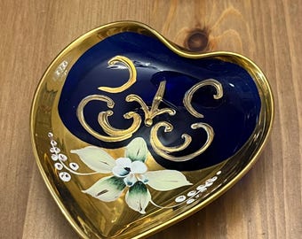 Peut inclure: Un plat en forme de cœur avec un intérieur bleu cobalt et un bord doré. Le plat est décoré de volutes peintes en or et d'une fleur blanche avec des feuilles vertes.