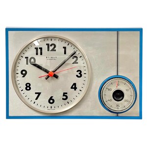 Peut inclure: Une horloge murale rectangulaire avec un cadre bleu et un cadran en argent brossé. Elle présente un grand cadran circulaire avec des chiffres noirs, des aiguilles noires pour les heures et les minutes, et une trotteuse rouge. Un minuteur est à droite.