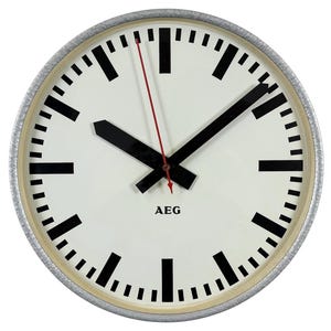 May include: A white clock face with black hour and minute markers and black hands. The clock has a silver rim and the word "AEG" printed in black at the bottom.