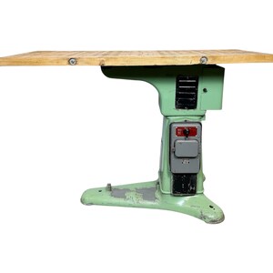 Könnte beinhalten: Ein vintage grüner Metalltisch mit einer Holzplatte. Der Tisch hat ein einzigartiges industrielles Design mit einem Sockel, der einer Maschine ähnelt. Der Tisch hat einen Schalter und eine Steckdose an der Seite.