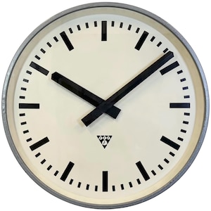 Può includere: Un grande quadrante di orologio rotondo e bianco con lancette delle ore e dei minuti nere. L'orologio ha indici orari in numeri romani neri e un triangolo nero con un piccolo orologio all'interno in basso. L'orologio è circondato da una cornice in metallo argentato.