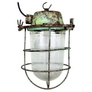 Puede incluir: Lámpara colgante industrial vintage con una pantalla de vidrio transparente y acanalado, encerrada en una jaula de metal desgastado. La lámpara tiene una parte superior pintada de verde y un aspecto oxidado y envejecido, lo que sugiere que es una pieza antigua o recuperada.