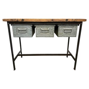 Op de afbeelding: Een rustieke consoletafel met een houten blad en drie metalen lades. De tafel heeft een zwart metalen frame en is ontworpen voor opslag en display. Het houten blad is lichtbruin.