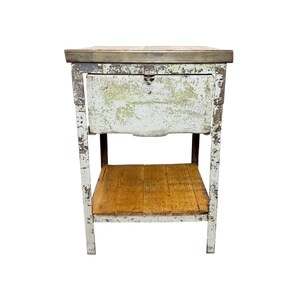 Peut inclure: Une table d'appoint carrée dotée d'un cadre en métal peint en blanc vieilli avec des signes d'usure visibles. La table comprend un tiroir supérieur unique avec une petite poignée en métal et une étagère inférieure en planches de bois clair.