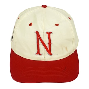 Puede incluir: Una gorra de béisbol blanca con una visera roja. La parte delantera de la gorra tiene una letra "N" bordada en rojo con un contorno blanco. La gorra tiene un pequeño símbolo "TM" blanco debajo de la letra.