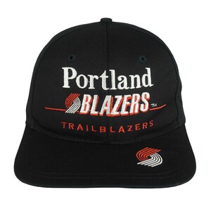 May include: A black baseball cap featuring the embroidered text Portland BLAZERS in white and red, with the word TRAILBLAZERS below it in red. A small red and white pinwheel logo is embroidered on the left side of the flat brim and the centre.