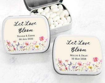 May include: Two small, silver, rectangular tins with floral designs and the text "Let Love Bloom". The top tin is open and contains white mints. The bottom tin is closed and has the text "Nicole & Eddie 06 Aug 2026" on the front. The top tin has the text "Millie & Dean 10 May 2028" on the front.