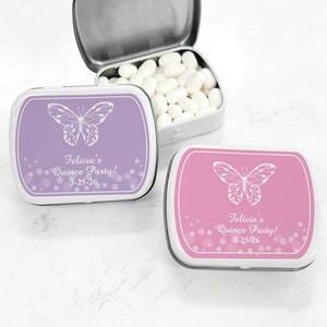May include: Two small, white metal tins with lids. The tins are decorated with a purple and pink butterfly design and the text "Felicia's Quince Party! 3-25-26".