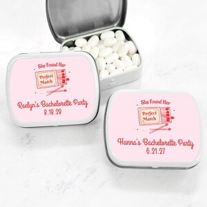 May include: Two pink and white mint tins with a red and white illustration of a matchbook and matches. The text on the tins reads "She Found Her Perfect Match" and includes the names of the brides-to-be and their bachelorette party dates.